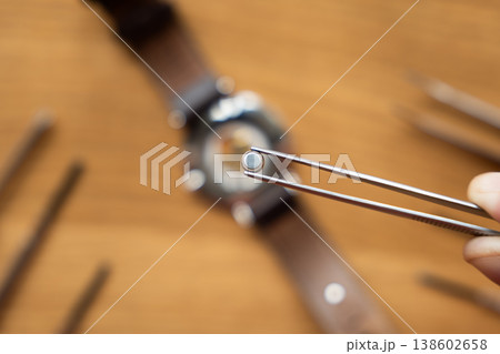 Close-up of tweezers holding a small watch battery above a wooden workbench with tools and a watch in the background Close-up of tweezers holding a small watch battery above a wooden workbench with tools and a watch in the background 138602658