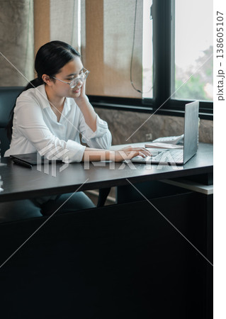 A focused woman in a modern office setting, working on a laptop with natural light streaming in through large windows. 138605107