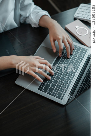 Close-up of hands typing on a laptop keyboard, surrounded by tech gadgets on a wooden desk in an office environment. 138605108