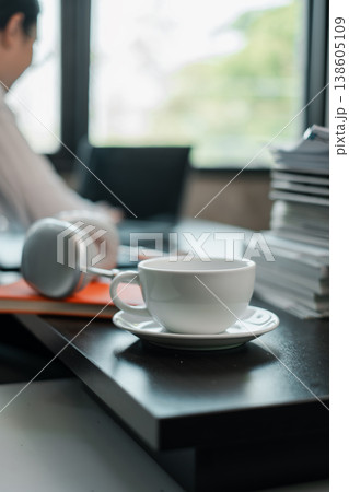A serene office setting featuring a coffee cup, headphones, and stacked documents on a desk by a window. 138605109