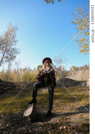 Beautiful blonde girl playing acoustic guitar and singing relaxing enjoying outdoors on sunny autumn day. Hippie woman in boho style learning and practicing playing musical instrument. Fall leaves 138609581
