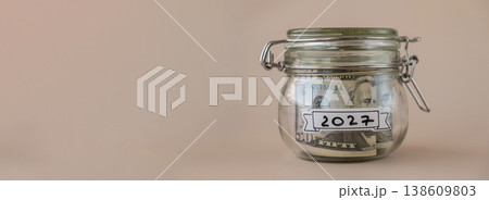Saving Money In Glass Jar filled with Dollars banknotes. 2027 year transcription in front of jar. Managing personal finances extra income for future insecurity Saving Money In Glass Jar filled with Dollars banknotes. 2027 year transcription in front of jar. Managing personal finances extra income for future insecurity 138609803
