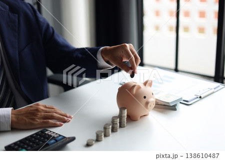 Man putting a coin into a piggy bank with stacks of coins and money on the table. Saving money and financial planning, financial growth, investment, and future planning concept. Man putting a coin into a piggy bank with stacks of coins and money on the table. Saving money and financial planning, financial growth, investment, and future planning concept. 138610487
