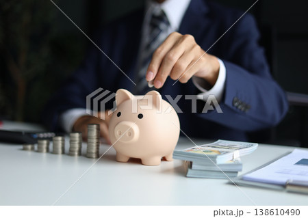 Man putting a coin into a piggy bank with stacks of coins and money on the table. Saving money and financial planning, financial growth, investment, and future planning concept. Man putting a coin into a piggy bank with stacks of coins and money on the table. Saving money and financial planning, financial growth, investment, and future planning concept. 138610490