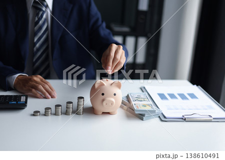 Man putting a coin into a piggy bank with stacks of coins and money on the table. Saving money and financial planning, financial growth, investment, and future planning concept. Man putting a coin into a piggy bank with stacks of coins and money on the table. Saving money and financial planning, financial growth, investment, and future planning concept. 138610491