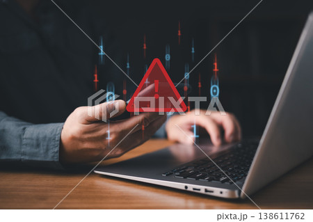 A person using a smartphone and laptop with a red warning alert icon, representing cybersecurity threat, malware attack, phishing, or data breach risk in the digital environment. 138611762