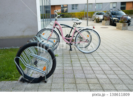 A white bicycle is secured to a metal bike rack on a tiled sidewalk. Colorful artwork adorns the nearby building, creating a vibrant urban atmosphere in the bright daylight. A white bicycle is secured to a metal bike rack on a tiled sidewalk. Colorful artwork adorns the nearby building, creating a vibrant urban atmosphere in the bright daylight. 138611809