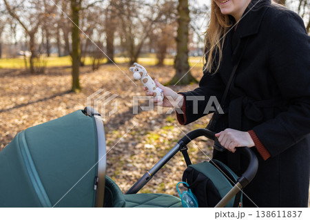 Mother holding baby toy while standing next to stroller in park, outdoor parenting and childcare lifestyle concept. Mother holding baby toy while standing next to stroller in park, outdoor parenting and childcare lifestyle concept. 138611837