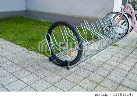 A white bicycle is secured to a metal bike rack on a tiled sidewalk. Colorful artwork adorns the nearby building, creating a vibrant urban atmosphere in the bright daylight. 138612143