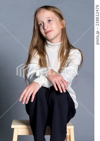 Girl sits wooden step stool with hands gently placed on her knee, subtle smile and quiet confidence 138612478