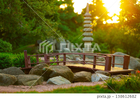 Serene Japanese garden scene with a small wooden bridge and stone pagoda at sunset surrounded by lush greenery Serene Japanese garden scene with a small wooden bridge and stone pagoda at sunset surrounded by lush greenery 138612682