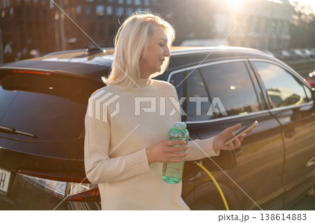 Eco conscious blonde woman using smartphone while standing next to electric vehicle during charging session. Eco conscious blonde woman using smartphone while standing next to electric vehicle during charging session. 138614883