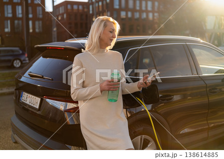 Electric vehicle plugged into charger at urban location while a woman checks her smartphone. Electric vehicle plugged into charger at urban location while a woman checks her smartphone. 138614885
