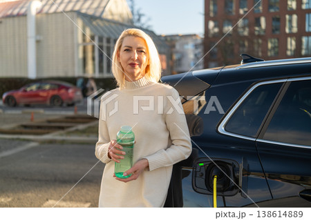 A woman stands next to an electric vehicle, drinking from a green reusable water bottle. The scene is set in an urban area with buildings and parked cars visible in the background. A woman stands next to an electric vehicle, drinking from a green reusable water bottle. The scene is set in an urban area with buildings and parked cars visible in the background. 138614889