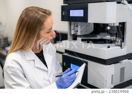 Laboratory technician taking notes while operating a sophisticated HPLC chromatography machine at research facility. Laboratory technician taking notes while operating a sophisticated HPLC chromatography machine at research facility. 138614908