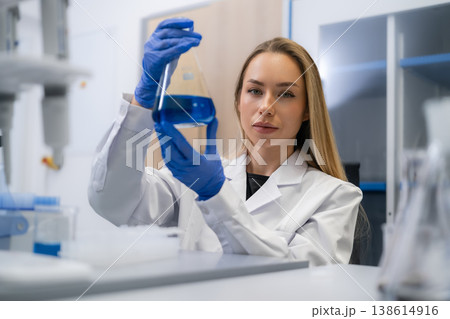 Analytical chemist assessing a blue liquid sample in an Erlenmeyer flask at laboratory workstation. Analytical chemist assessing a blue liquid sample in an Erlenmeyer flask at laboratory workstation. 138614916