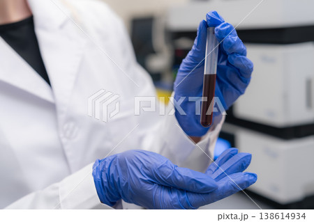 Laboratory technician examines blood sample held in test tube at clinical workstation. Clean lab setting with advanced analytical equipment visible in background. Laboratory technician examines blood sample held in test tube at clinical workstation. Clean lab setting with advanced analytical equipment visible in background. 138614934