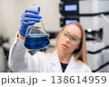 Laboratory technician inspecting blue colored liquid in Erlenmeyer flask while wearing protective gloves and eyewear. 138614959