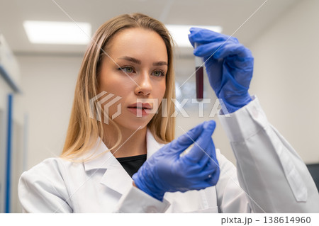 Laboratory technician analyzing a blood sample contained within a test tube at a clinical research facility. Clean, well lit environment with sterile surfaces and laboratory equipment visible. 138614960