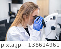 Technician examining samples through high resolution microscope in clinical laboratory. Clean workspace with modern instruments and digital displays visible in the background. 138614961