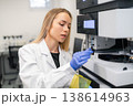 Technician configuring advanced HPLC liquid chromatography system at pharmaceutical laboratory station. Clean workspace with digital readout and safety gear visible in the background. 138614963