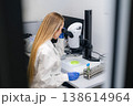 Scientist examining biological specimens through high powered microscope at laboratory workstation. Bright workspace featuring sample dishes and test tubes with colored liquids is visible. 138614964