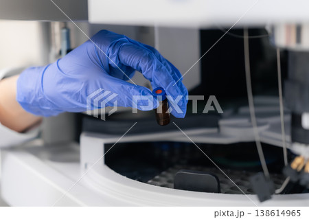 Technician operating automated liquid handling system HPLC, inserting an amber vial with a blue cap into the analyzer. Clean laboratory environment featuring precision equipment and organized Technician operating automated liquid handling system HPLC, inserting an amber vial with a blue cap into the analyzer. Clean laboratory environment featuring precision equipment and organized 138614965