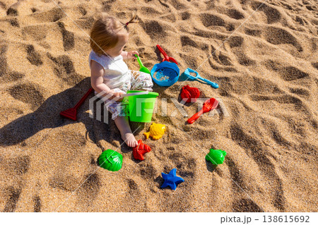 Toddler sitting on sandy beach playing with plastic bucket shovel and sand molds. Outdoor beach photography. Childhood and summer vacation concept. Design for poster, banner, postcard. Top view shot 138615692