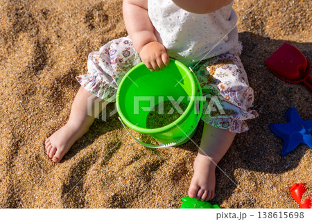 Toddler sitting on sandy beach holding green plastic bucket with sand toys around. Outdoor beach photography. Childhood and summer vacation concept. Design for poster, banner, postcard. Top view shot Toddler sitting on sandy beach holding green plastic bucket with sand toys around. Outdoor beach photography. Childhood and summer vacation concept. Design for poster, banner, postcard. Top view shot 138615698