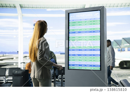 Female business consultant reading flight information on screen in airport, preparing for business trip departure in busy terminal waiting area. Travelers near the boarding gate. Female business consultant reading flight information on screen in airport, preparing for business trip departure in busy terminal waiting area. Travelers near the boarding gate. 138615810