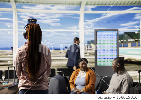 Teen listens to music, black girl with headphones, waiting for vacation flight, busy departure area at the airport terminal. Solo traveler waiting impatiently for boarding plane in lounge. 138615816