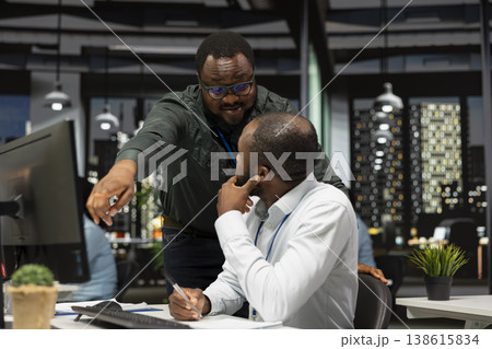 African american colleagues team in office reviewing reports on computer monitor, focusing on report writing and project planning to drive results and success. Doing overtime together. 138615834