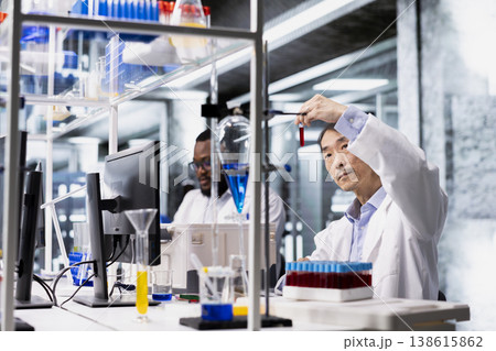 Senior biotechnologist in lab looking at blood sample in test tubes. Asian man in modern laboratory workspace analyzing sanguine fluid specimen in vial used for medical research 138615862
