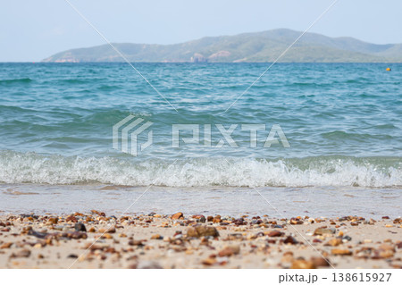 Clear blue ocean waves softly break on a sandy shore scattered with pebbles, with a calm island landscape in the distance under bright daylight, creating a peaceful coastal scene. 138615927