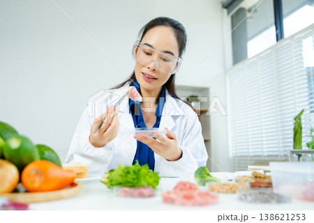 Female scientist in lab coat and goggles examining raw meat with tweezers in food lab. Nutrition, protein, food safety, and research concept. Female scientist in lab coat and goggles examining raw meat with tweezers in food lab. Nutrition, protein, food safety, and research concept. 138621253