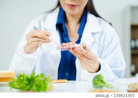 Female scientist in lab coat and goggles examining raw meat with tweezers in food lab. Nutrition, protein, food safety, and research concept. 138621303