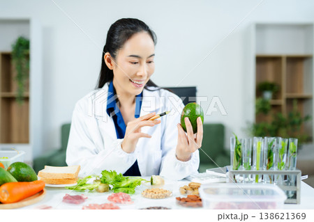 Female scientist in lab coat holding avocado while analyzing healthy ingredients. Nutrition science, food safety, and clean eating research concept. Female scientist in lab coat holding avocado while analyzing healthy ingredients. Nutrition science, food safety, and clean eating research concept. 138621369