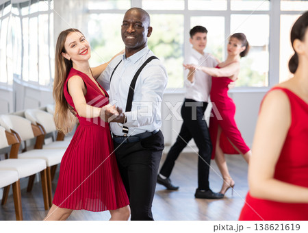 Positive woman in red dress and African american man practicing waltz dance moves in pair during group party in dancing studio Positive woman in red dress and African american man practicing waltz dance moves in pair during group party in dancing studio 138621619