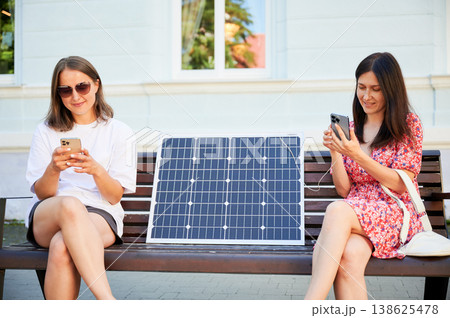 Two women sit on bench, using smartphone connected to solar panel. Integration of sustainable renewable energy into everyday life, demonstrating practical use of solar power for charging devices. 138625478