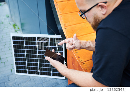 Bearded man using smartphone connected to photovoltaic solar panel. Integration of sustainable renewable energy into everyday life, demonstrating practical use of solar power for charging devices. 138625484