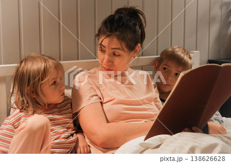 Family bedtime stories. A mother reads a story to her children, wrapped in a blanket. Children listen to soothing stories. A cozy family scene before bed in a softly lit bedroom. Copy space. Banner Family bedtime stories. A mother reads a story to her children, wrapped in a blanket. Children listen to soothing stories. A cozy family scene before bed in a softly lit bedroom. Copy space. Banner 138626628