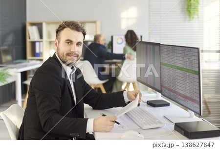 Smiling accountant at desk with spreadsheets on dual monitors in bright office 138627847