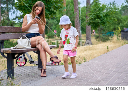 Mother reads message while relaxing on bench with her daughter Mother reads message while relaxing on bench with her daughter 138630236