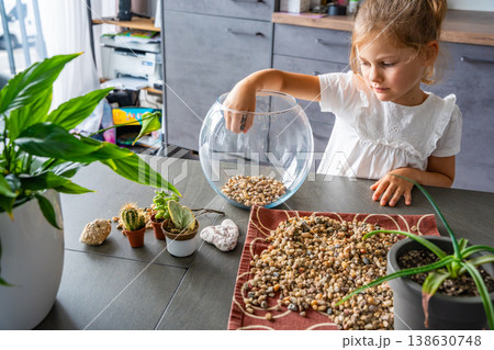 Little girl pouring stones into glass container to create drainage for future florarium or closed ecosystem. Hands-on learning, eco-friendly creativity and early involvement in nature-based projects. 138630748