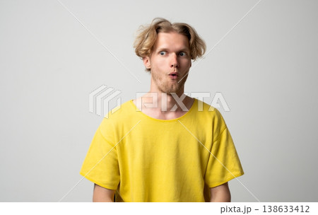 Young blond man in a yellow t-shirt expressing shock and surprise, widening eyes and mouth open on a light gray background, realizing unexpected news or a sudden event Young blond man in a yellow t-shirt expressing shock and surprise, widening eyes and mouth open on a light gray background, realizing unexpected news or a sudden event 138633412