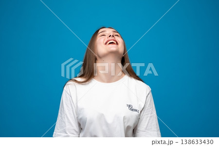 Young woman smiling and laughing looking up with joy, expressing happiness and cheerful emotions on a blue background Young woman smiling and laughing looking up with joy, expressing happiness and cheerful emotions on a blue background 138633430