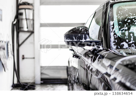Car wash, black car covered foam at indoor station. Side view with mirror and body panels in soap, cleaning process, glossy surface care, automotive washing and detailing service Car wash, black car covered foam at indoor station. Side view with mirror and body panels in soap, cleaning process, glossy surface care, automotive washing and detailing service 138634654