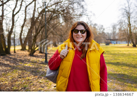 Happy woman in yellow vest and sunglasses walking along park path with backpack, enjoying sunny day and outdoor lifestyle. Happy woman in yellow vest and sunglasses walking along park path with backpack, enjoying sunny day and outdoor lifestyle. 138634667