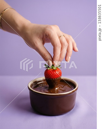 Hand Dipping Fresh Strawberry into Melted Glossy Chocolate Isolated on a Solid Soft Lavender Background 138635501
