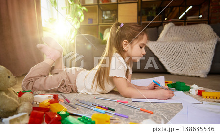 Child lying on floor drawing with pencil surrounded by toys and papers. Child lying on floor drawing with pencil surrounded by toys and papers. 138635536